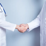 Deal! Concept of collaboration in medicine. Close up photo of two doctors' handshake on gray background